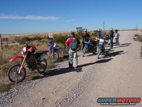 Several dirt bikes and riders stopped on a dusty road in a dry, open area