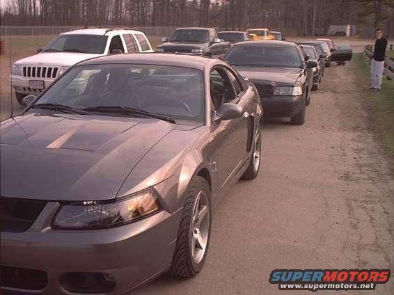 Gray Ford Mustang in foreground, line of cars behind it. Person standing on right