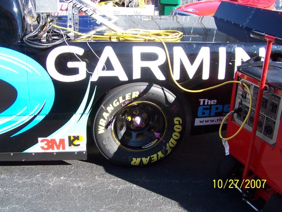 Side view of a black NASCAR race car with large white 'GARMIN' lettering and a Goodyear tire. A red generator is connected by a yellow cable
