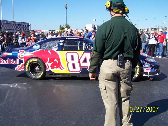 Red Bull #84 NASCAR race car, side view, with crowd and official in foreground