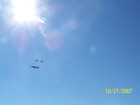 Blimp and two fighter jets flying in a clear blue sky with the sun shining
