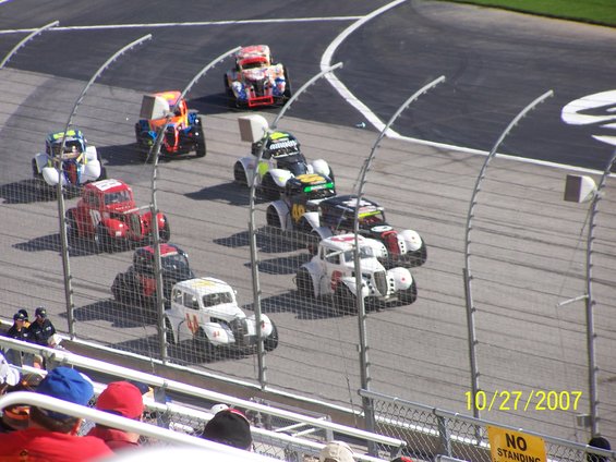 several stock cars racing on a track, some with colorful paint jobs, on a sunny day