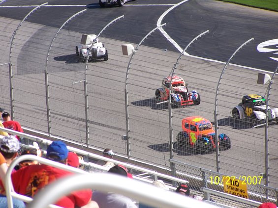 Stock cars racing on a track, one with orange and blue livery, another with red and white