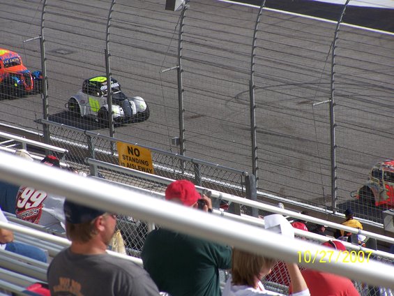 Stock cars racing on a track, viewed from the stands. A sign reads 'NO STANDING IN AISLEWAY'