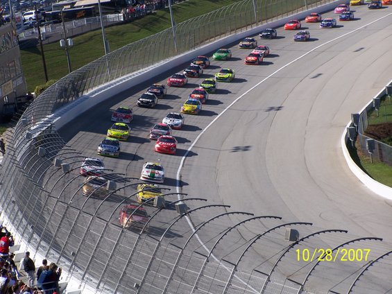 Multiple stock cars racing on a track, with spectators in the background