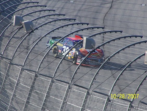 Stock car with number 32 on the side races on a track, seen from above behind a fence