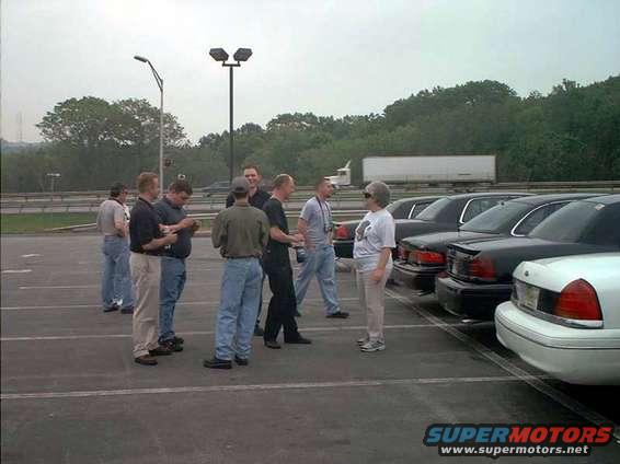 Several guys stand around in a parking lot, talking near a line of parked sedans
