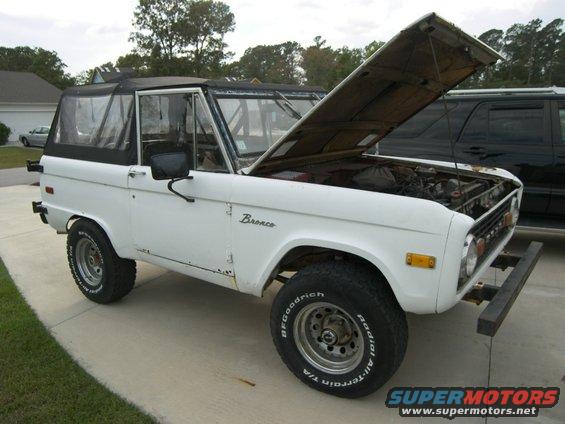 White 1974 Ford Bronco with black soft top, hood open, showing engine. Visible rust on lower body panels