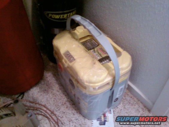 Yellow and gray car battery with a gray handle, wrapped in plastic. Part of a black tank with 'POWER' visible is behind it