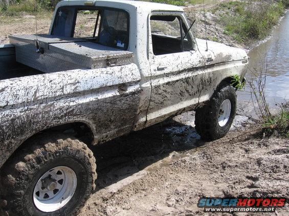 White 1977 Ford F-150 covered in mud, driving through a muddy track next to water