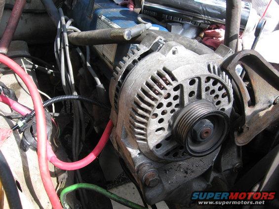 Alternator with red and green wires in a cluttered engine bay. Text 'SUPERMOTORS' visible