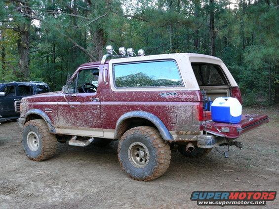 Muddy maroon 1988 Ford Bronco with off-road tires, tailgate down, cooler in back. Another dark SUV partially visible