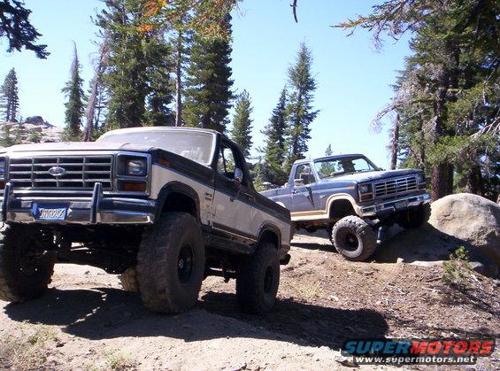 Two lifted Ford trucks, one Bronco and one pickup, on a dirt trail with trees