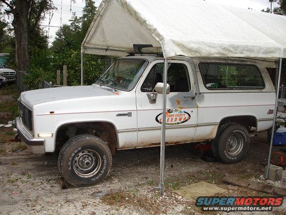 White GMC K5 Blazer with a canopy, parked under a tent. It has a 'Putnam County Speedway' decal on the door