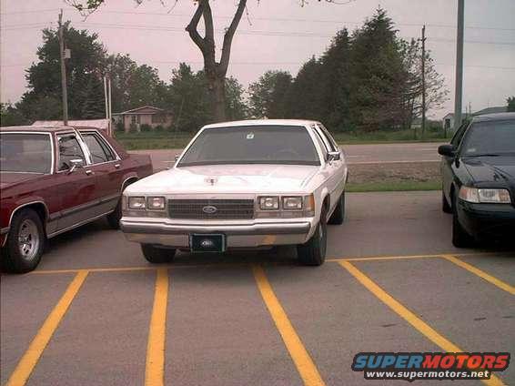 White Ford Crown Victoria parked in a lot, front view. The car has a chrome grille and rectangular headlights