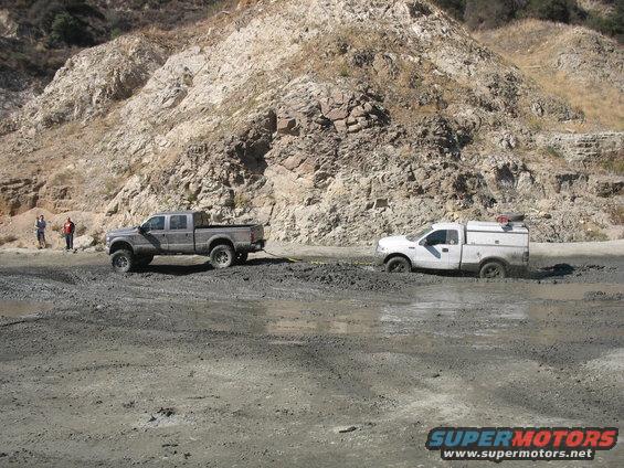 Gray pickup truck towing a white pickup truck stuck in mud near a rocky hill