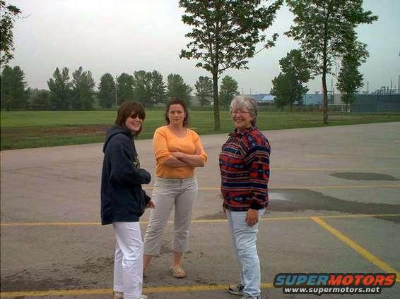 Three women stand in a parking lot with trees and a fence in the background