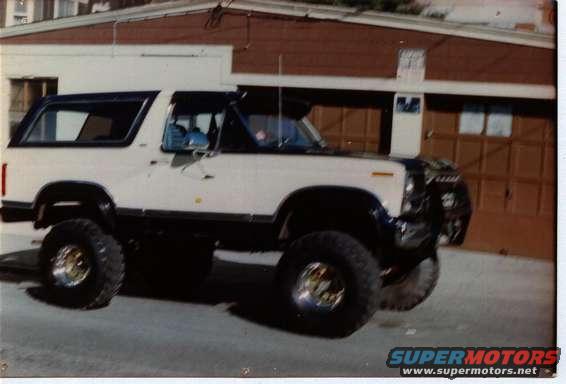 1980 Ford Bronco, white and black, lifted with large tires, parked in front of a garage
