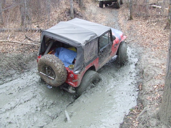 Red Jeep Wrangler with soft top stuck in deep mud on a wooded trail. Another vehicle is visible further up the trail
