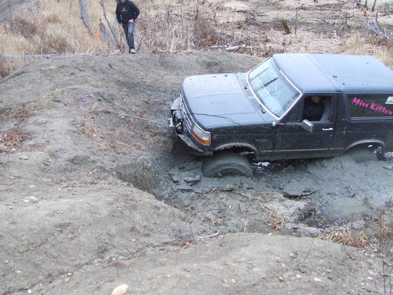 Black Ford Bronco stuck in a mud pit, with 'Miss Kitten' written on the side. A person stands on a dirt mound nearby