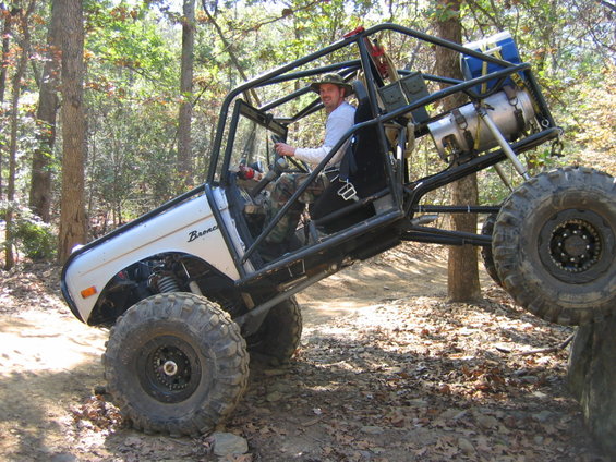 Off-road buggy with a roll cage and large tires climbing over a rock. Driver is wearing a hat