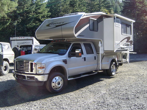 Silver Ford F-series truck with a Host camper shell, front 3/4 view, gravel lot, trees in background