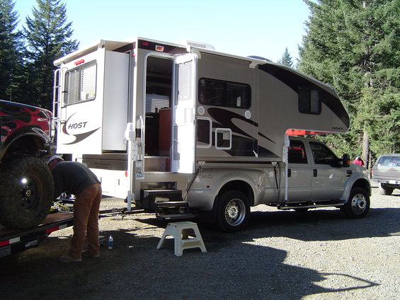 Silver Ford F-series truck with a Host camper shell, parked next to a trailer with a black vehicle. Trees and gravel ground