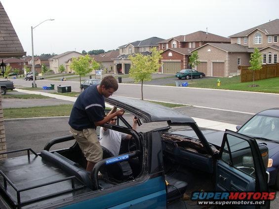Teal Ford Bronco with removed top, two men working on interior, parked in suburban driveway
