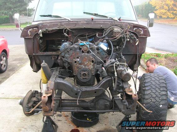 Brown truck chassis with engine removed, front suspension disassembled, on jack stands