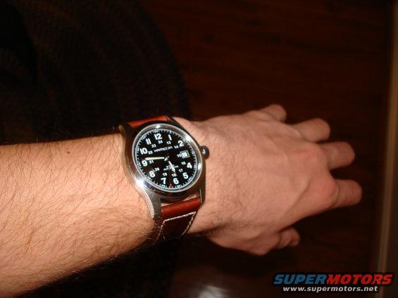 Watch with brown leather strap on a hairy arm. Black dial shows white numbers and hands