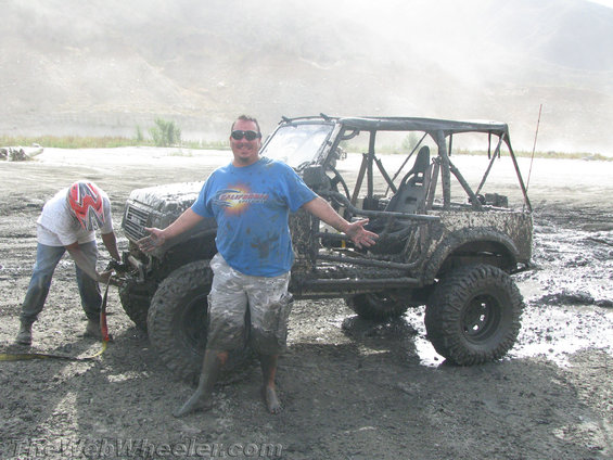 Mud-covered 1987 Suzuki Samurai with roll cage, off-road tires, and two people standing nearby