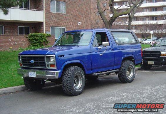 Blue Ford Bronco parked on street, next to a black SUV. Building in background