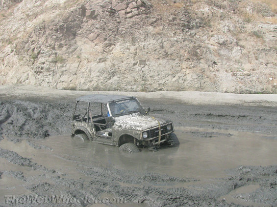Mud-covered Suzuki Samurai stuck in mud, front 3/4 view, soft top removed, roll cage visible