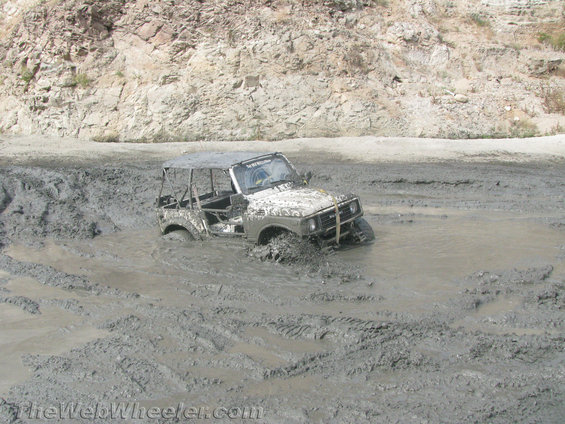 White 1987 Suzuki Samurai stuck in deep mud, front right tire submerged, off-road tires visible, roll cage present