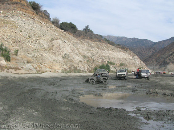Off-road vehicles navigating a muddy, dry riverbed with steep, rocky hillsides