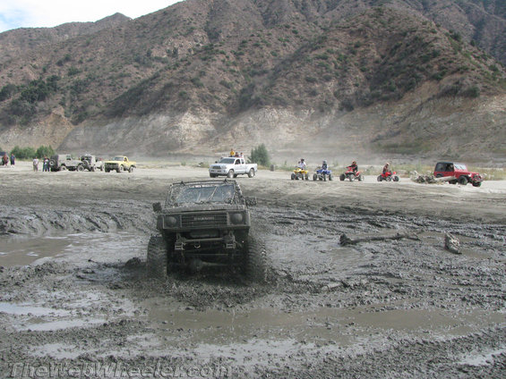 Mud-covered Suzuki Samurai driving through a muddy puddle, front view. Other vehicles and ATVs are visible in the background