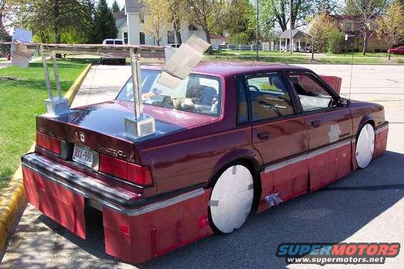 Maroon sedan with a large rear spoiler, red body kit, and white wheel covers. Rear view