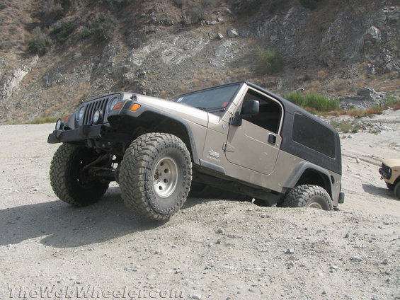 Tan Jeep Wrangler with a black hardtop is partially submerged in sand, front wheel turned