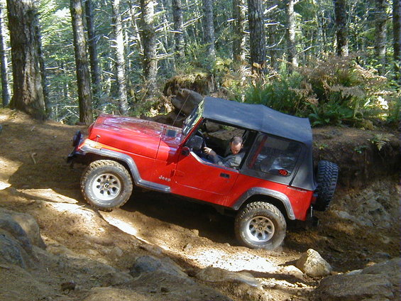Red Jeep Wrangler with a black soft top driving on a rocky, wooded trail