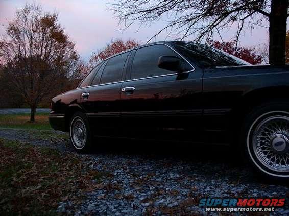 Black 1997 Ford Crown Victoria sedan, side view, parked on gravel with fall foliage