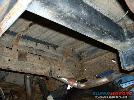 Underside of vehicle frame showing rust, suspension components, and exhaust pipe. Some text visible on tire