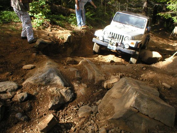 Silver 2005 Jeep Wrangler navigating a difficult rocky off-road trail with two people standing nearby