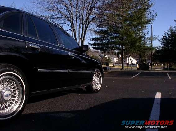 Side view of a black 1997 Ford Crown Victoria parked in a lot with trees and houses in the background