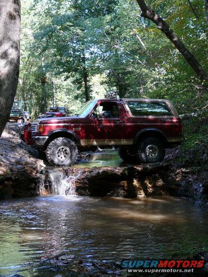 Red Ford Bronco driving through a stream in a wooded area. Another SUV is visible in the background