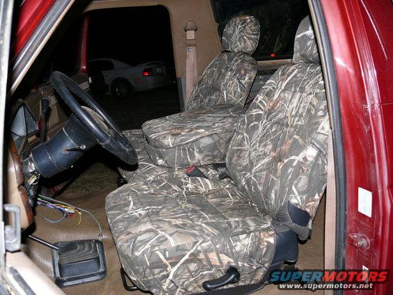Interior of a red vehicle with camouflage seat covers, steering wheel, and exposed wires. A white car is visible outside