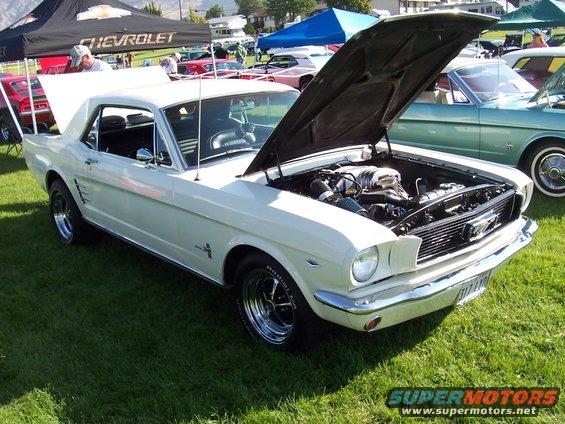 White 1965 Ford Mustang coupe with hood open, showing engine, parked on grass at a car show