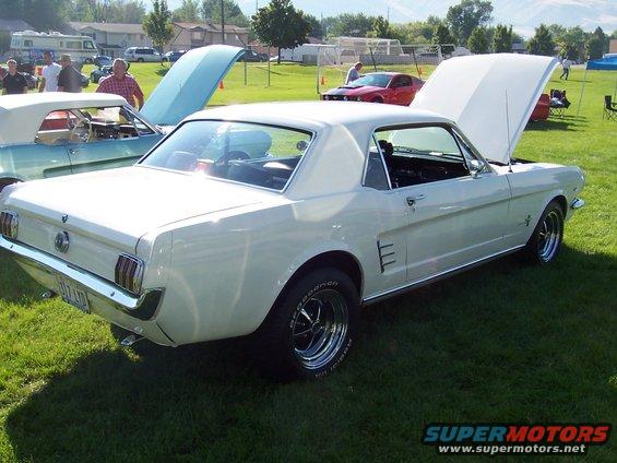 White 1965 Ford Mustang coupe parked on grass, hood open. Another classic car is visible to the left