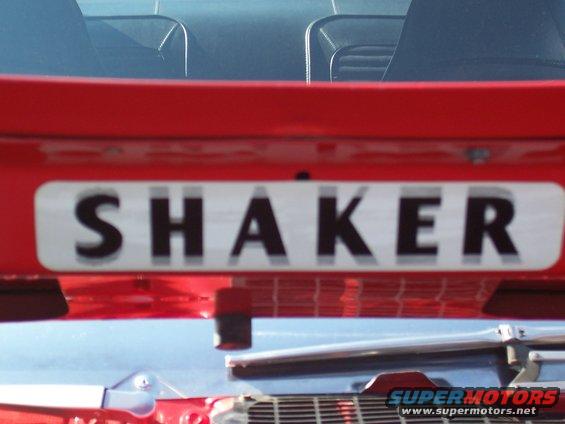 Red car's rear decklid with a "SHAKER" badge and part of a chrome bumper