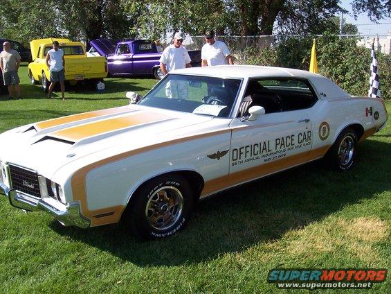 White Oldsmobile Cutlass Supreme with gold stripes, marked as official pace car for 56th annual Indianapolis 500 mile race on May 27, 1972. Yellow and purple pickup trucks are parked behind it