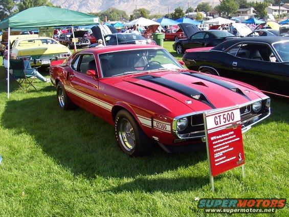 Red 1970 Shelby Mustang GT500 with black stripes and GT500 side decals, parked on grass at a car show